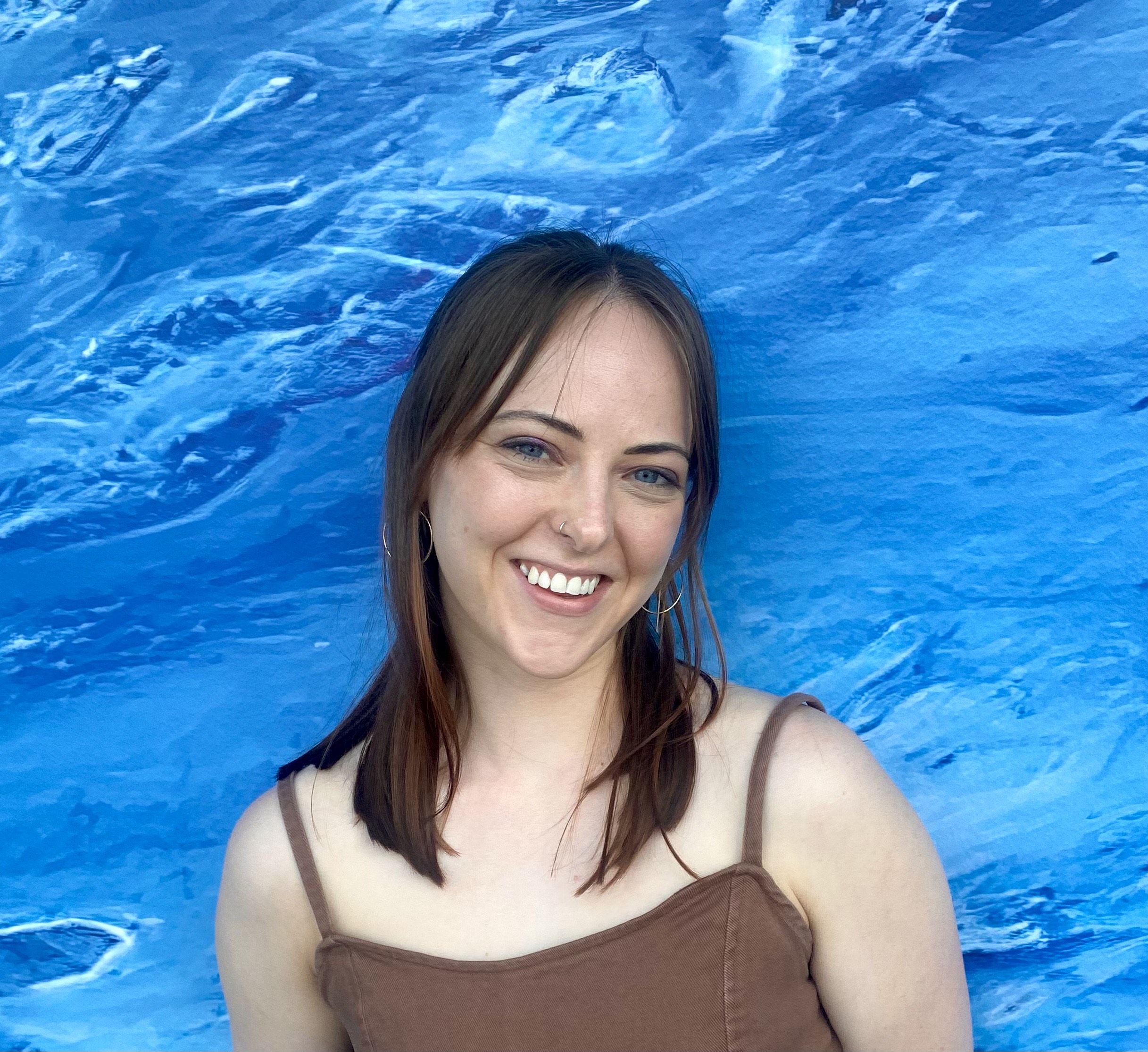A medium-haired white woman with blue eyes smiles in front of an oceanic backdrop